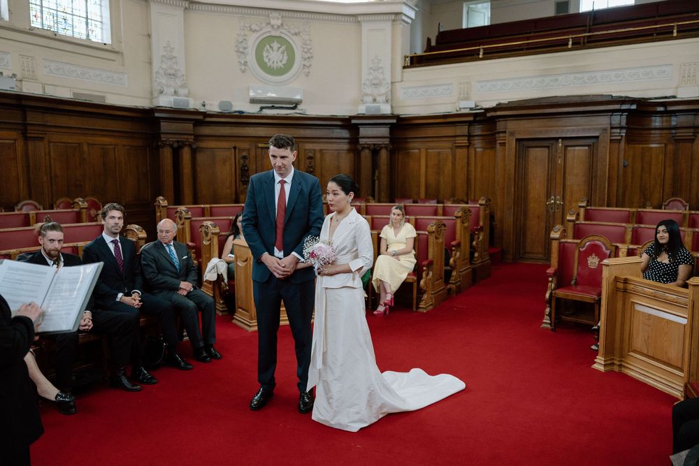 Couple exchanging vows at a formal wedding ceremony in an elegant wood-paneled hall with guests seated in red pews.