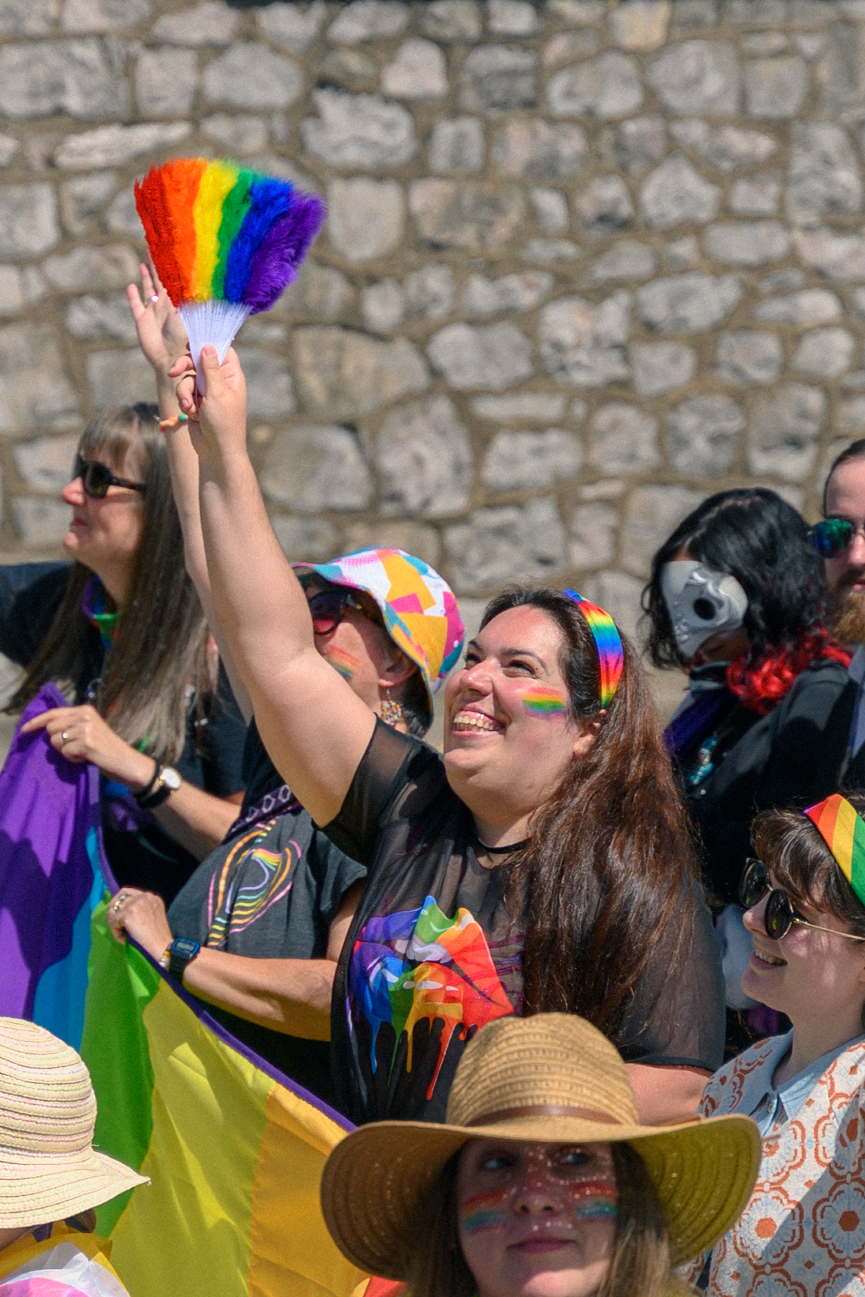 Woman raising rainbow feather fan at Pride parade, smiling crowd with colorful flags and face paint in background.