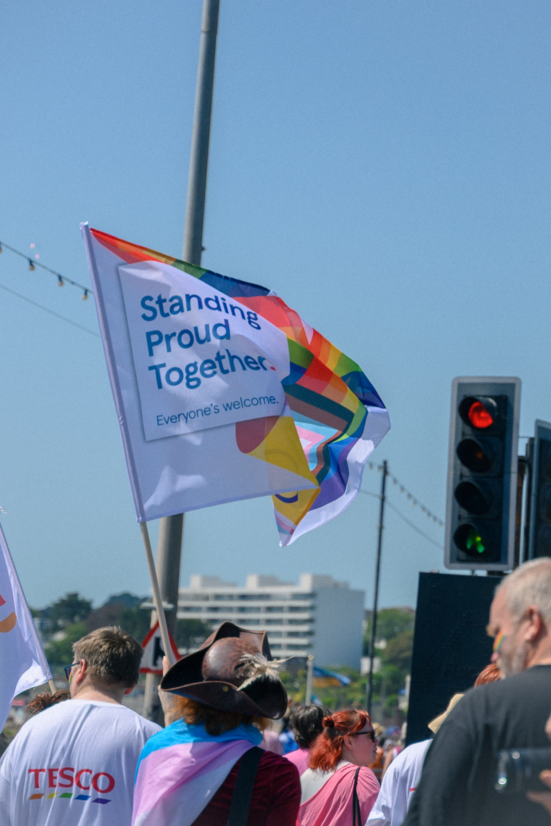 A Pride parade crowd waves a rainbow flag reading Standing Proud Together, Everyones welcome, with a Tesco shirt visible below.
