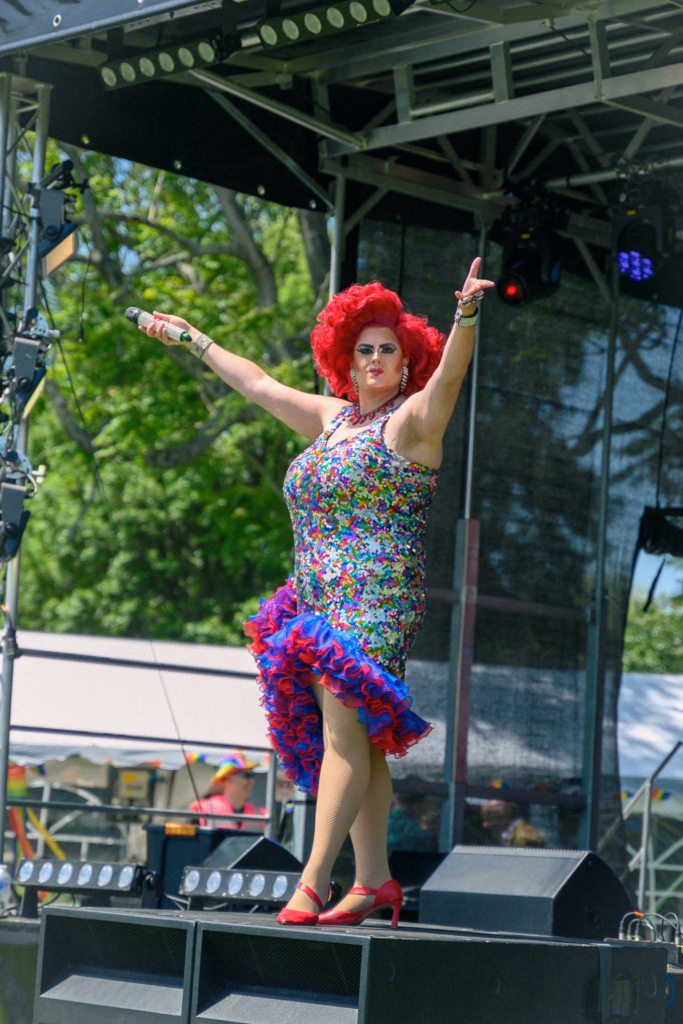 Drag performer in colorful dress and red wig dancing on outdoor stage at Pride festival.