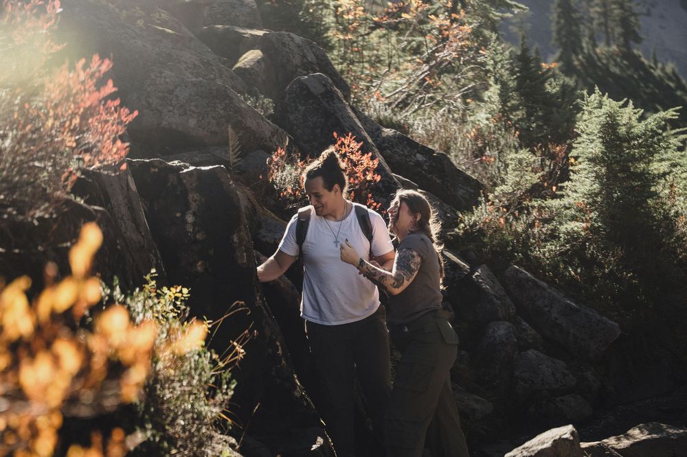 Two hikers pause on a rocky mountain trail surrounded by autumn foliage and evergreen trees in warm, golden sunlight.