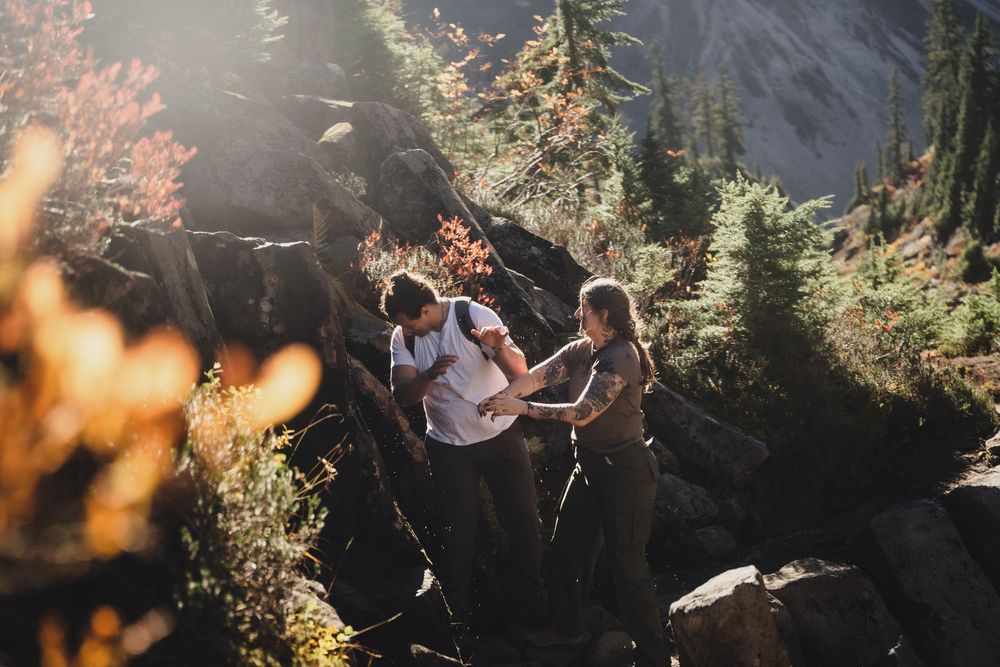 Two hikers hold hands while climbing a rocky mountain trail surrounded by evergreen trees and glowing autumn foliage.
