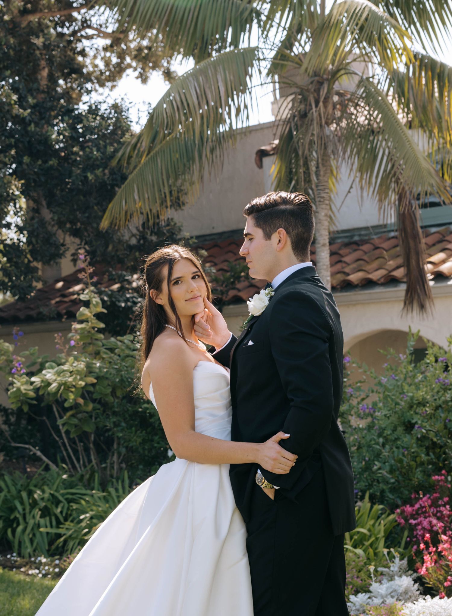 A bride in a strapless white gown stands close to her groom in a black suit near palm trees and flowering garden plants.