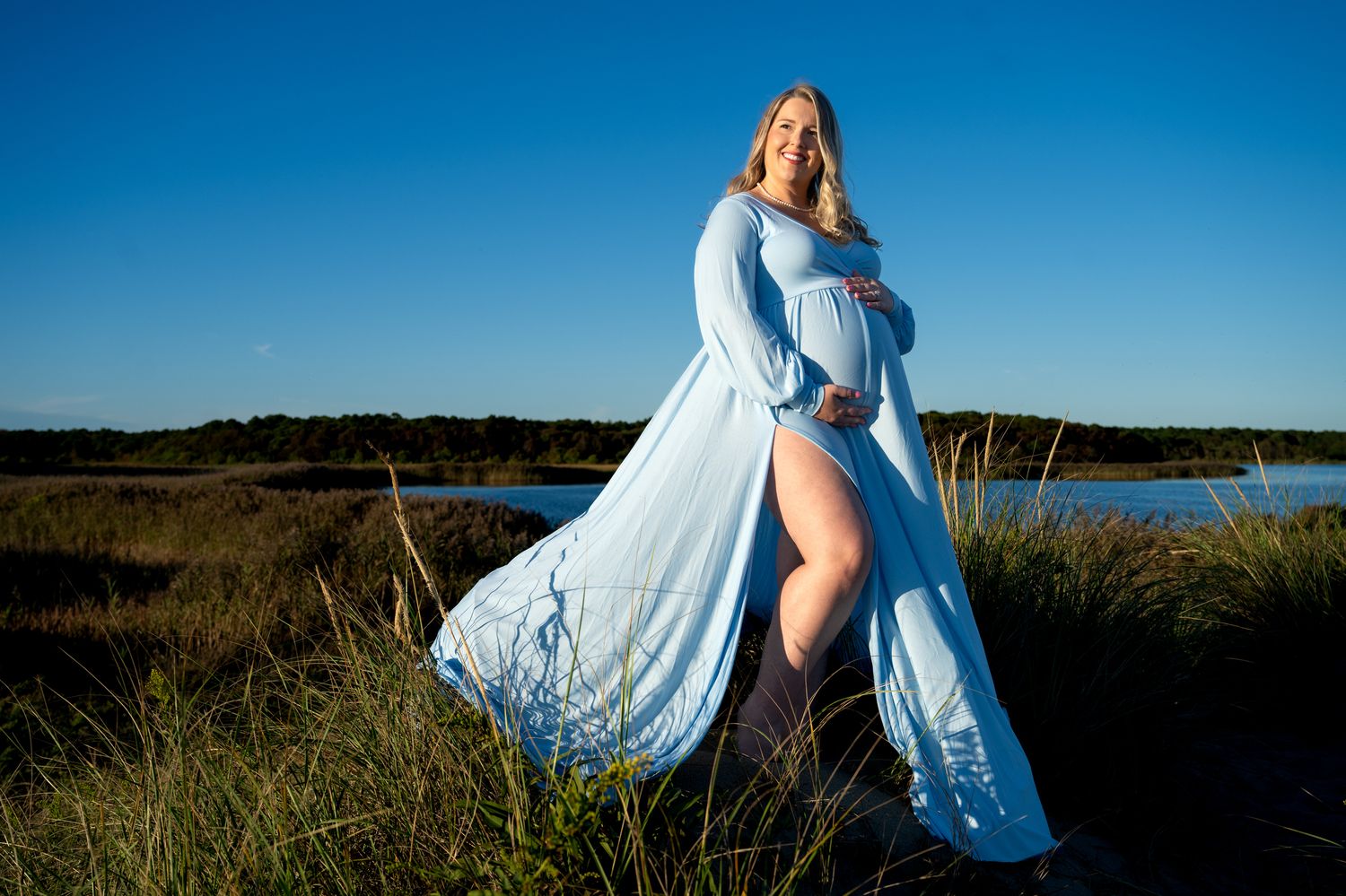 Pregnant woman in a flowing light blue maternity gown poses outdoors among tall grass beside calm water under a clear blue sky at sunset.