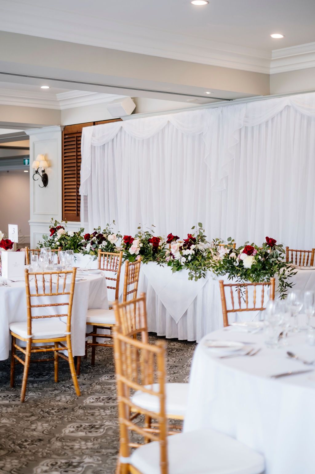 Elegant wedding reception hall with white draped backdrop, red and white floral garland, round tables & gold chiavari chairs Stamford Plaza