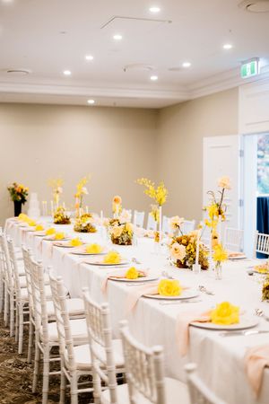 Long banquet table with white linens, yellow floral centerpieces, folded napkins and white chiavari chairs in an elegant event room Brisbane
