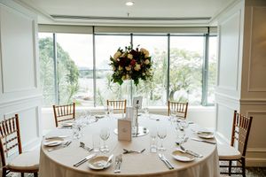 Elegant round banquet table set with white linens, glassware and a floral centerpiece, beside tall windows with scenic waterfront views QLD