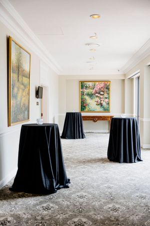 Elegant hotel corridor with three black-draped cocktail tables, ornate patterned carpet, and colorful floral paintings on cream walls QLD