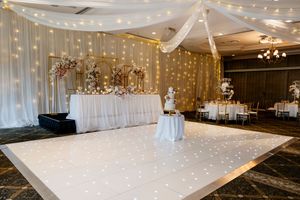 Elegant wedding reception room with a lit white dance floor, floral-decked head table, tiered cake and draped ceiling with fairy lights QLD