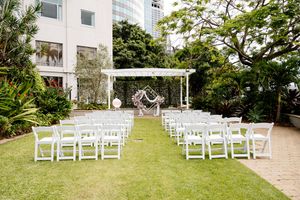 White folding chairs line a grassy aisle leading to a floral-decorated pergola at an outdoor garden wedding ceremony venue Stamford Plaza