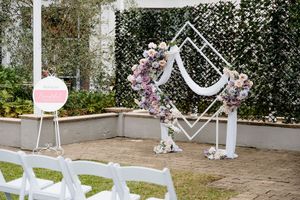 Outdoor wedding ceremony setup with a geometric arch draped in white fabric and pink floral arrangements, white chairs, and a welcome sign
