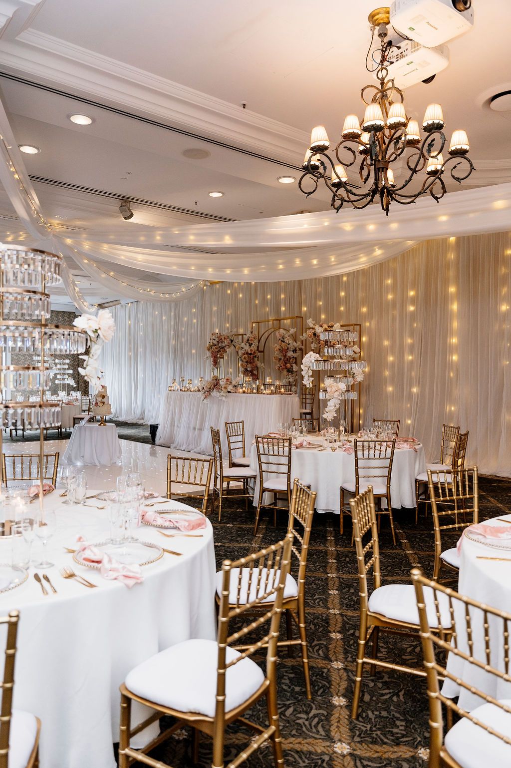 Elegant wedding reception hall with round tables, gold chiavari chairs, pink napkins, a black chandelier, and draped fairy-light curtains.