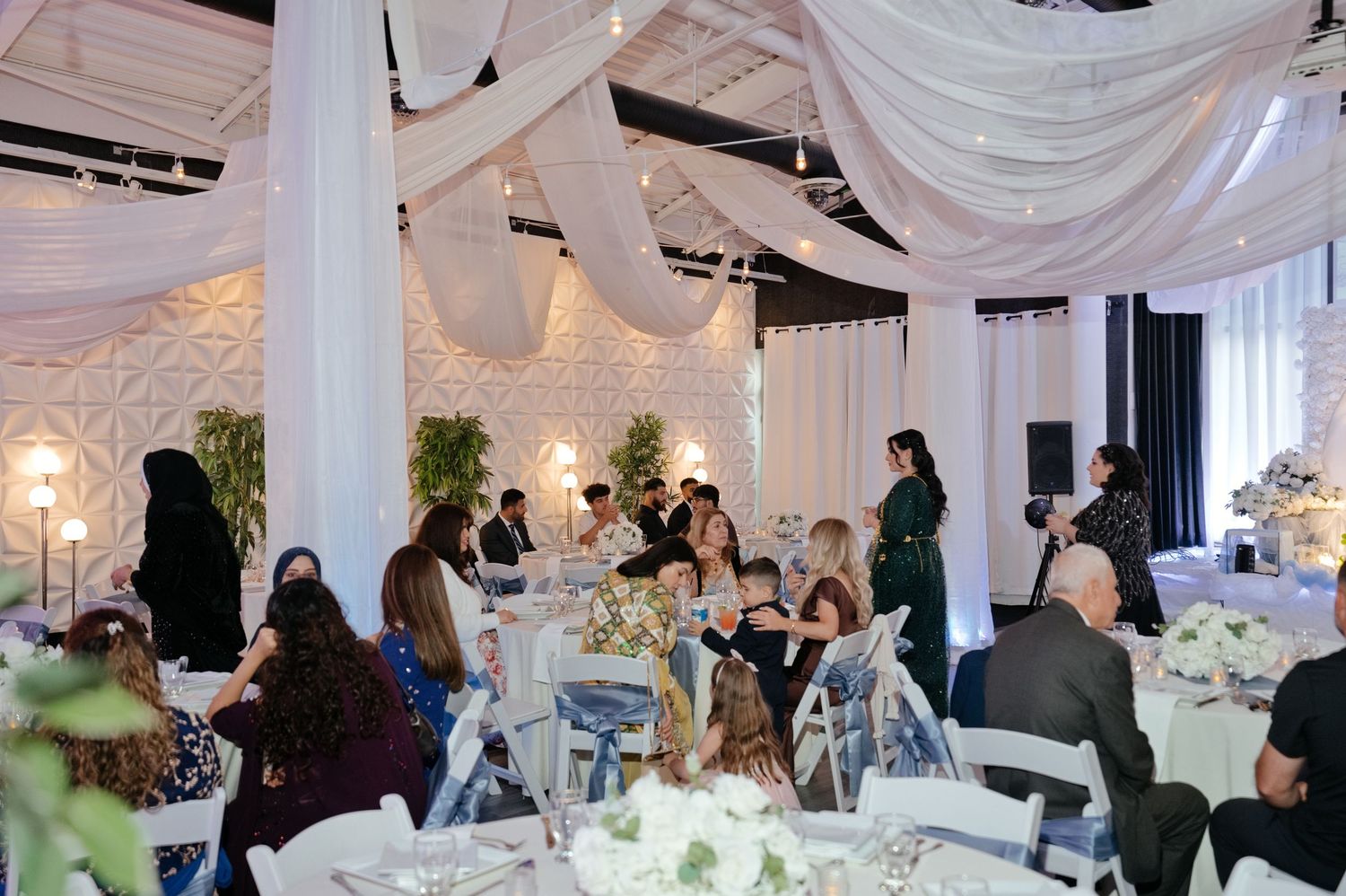 Wedding reception guests seated at white-linen tables beneath flowing draped ceiling fabric, with floral centerpieces and elegant decor.