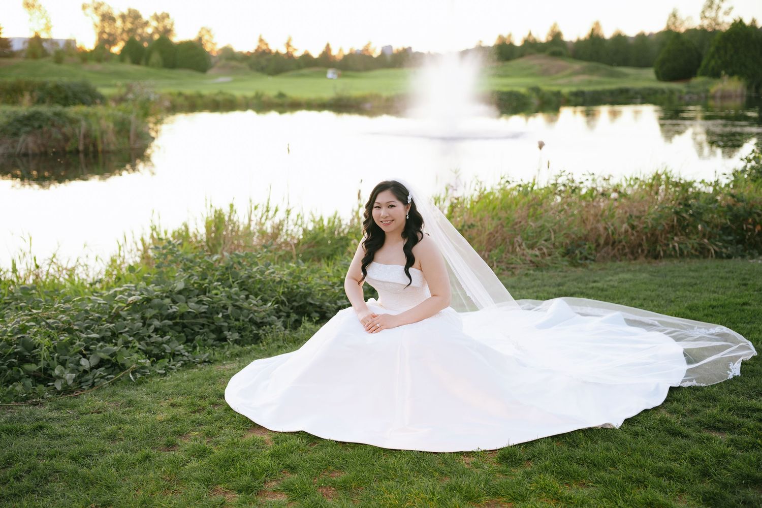 Bride in a flowing white wedding gown and veil sitting on a green lawn beside a tranquil pond with a fountain at sunset.