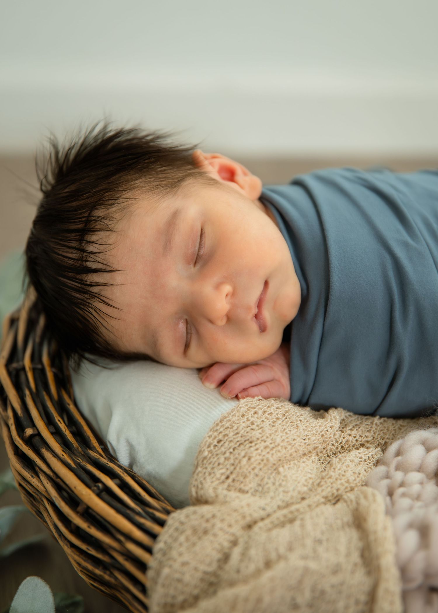 Peaceful newborn baby swaddled in blue wrap sleeping in a wicker basket with soft blankets.
