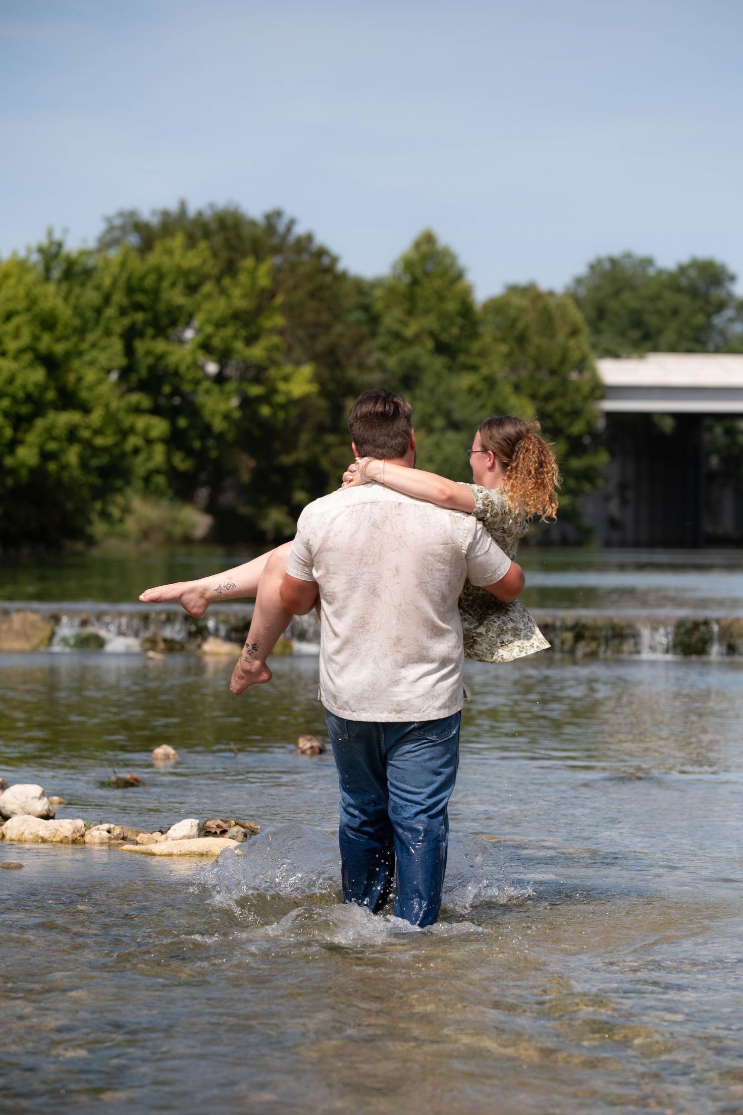 Two people wading in a shallow river, reaching toward each other, surrounded by lush green trees.