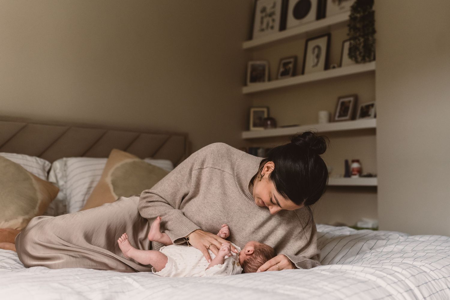 Mother tenderly leaning over her newborn baby lying on a white bed in a cozy bedroom.
