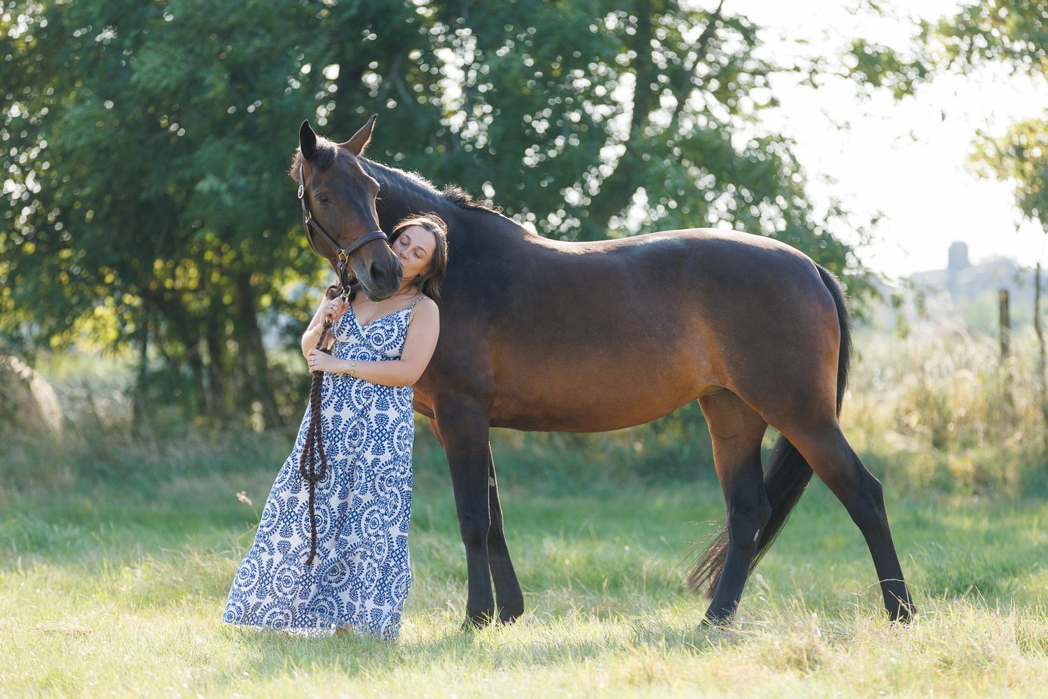 A woman sits on a log beside a brown horse in a field, holding its reins in a serene black and white photo.