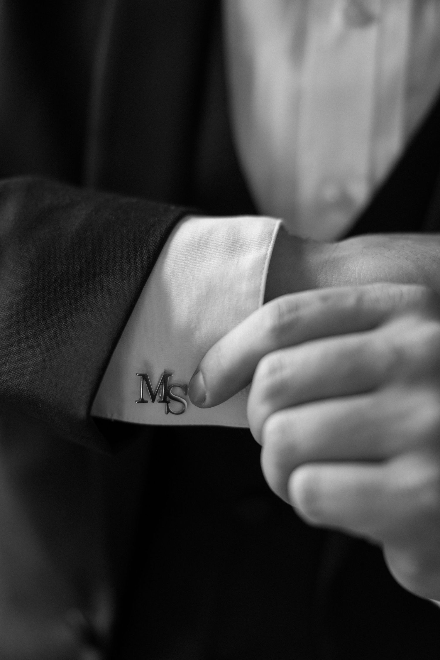Black and white photo of a man adjusting monogrammed MS cufflinks on a formal suit shirt sleeve.