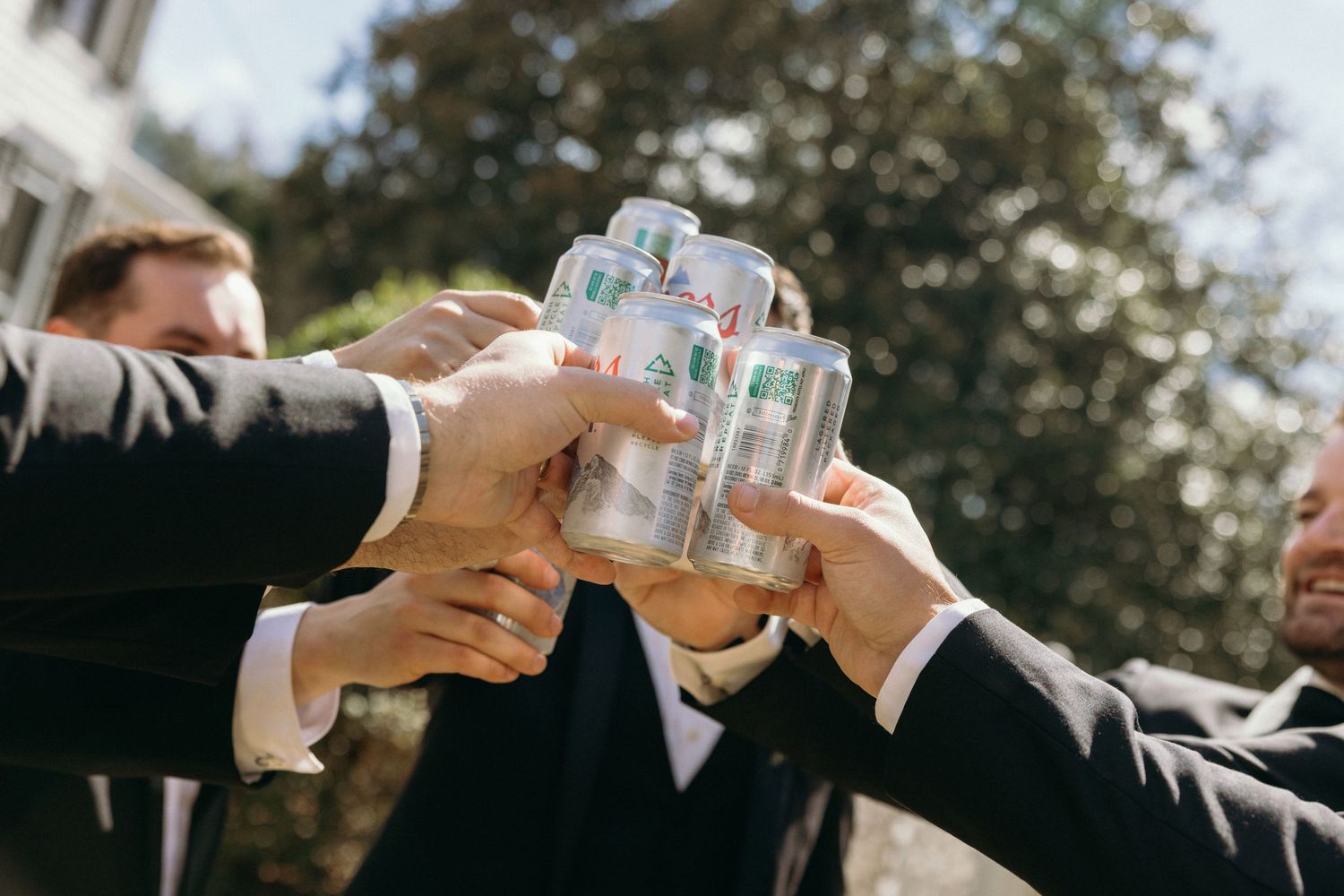 Groomsmen in black suits toasting with canned drinks outdoors at a wedding celebration in winter.