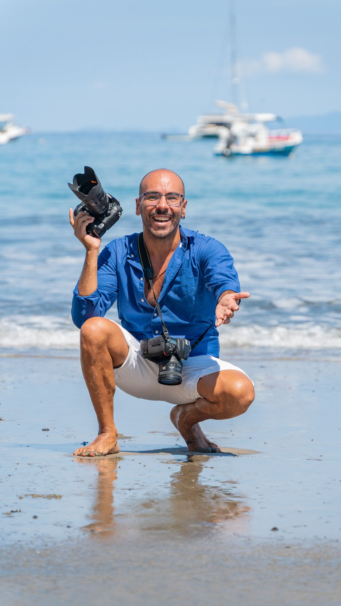 Smiling bald man in blue shirt crouching on beach holding camera with sailboats in background.