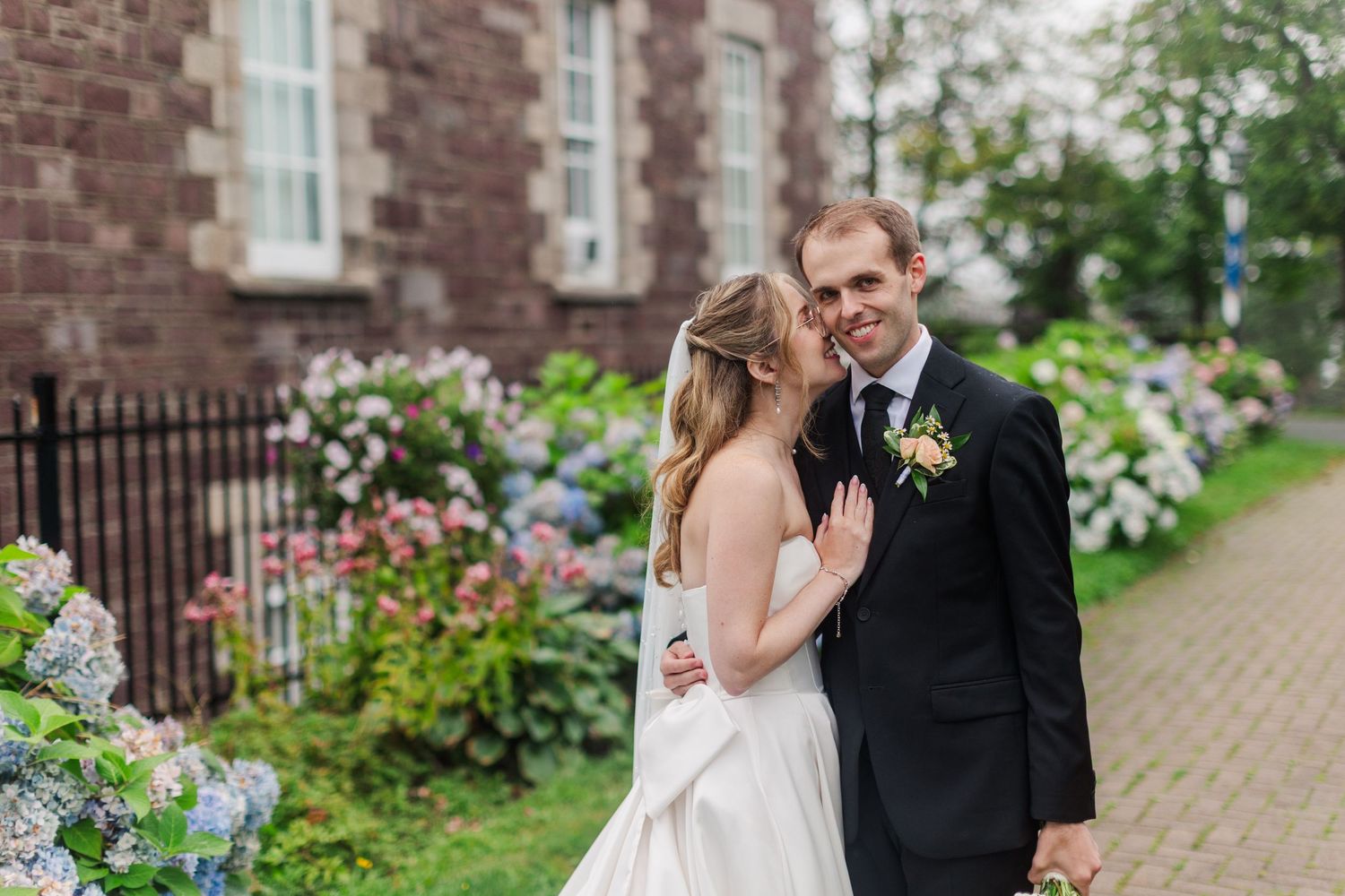 Bride and groom embrace near blooming hydrangeas outside a historic stone building on their wedding day.