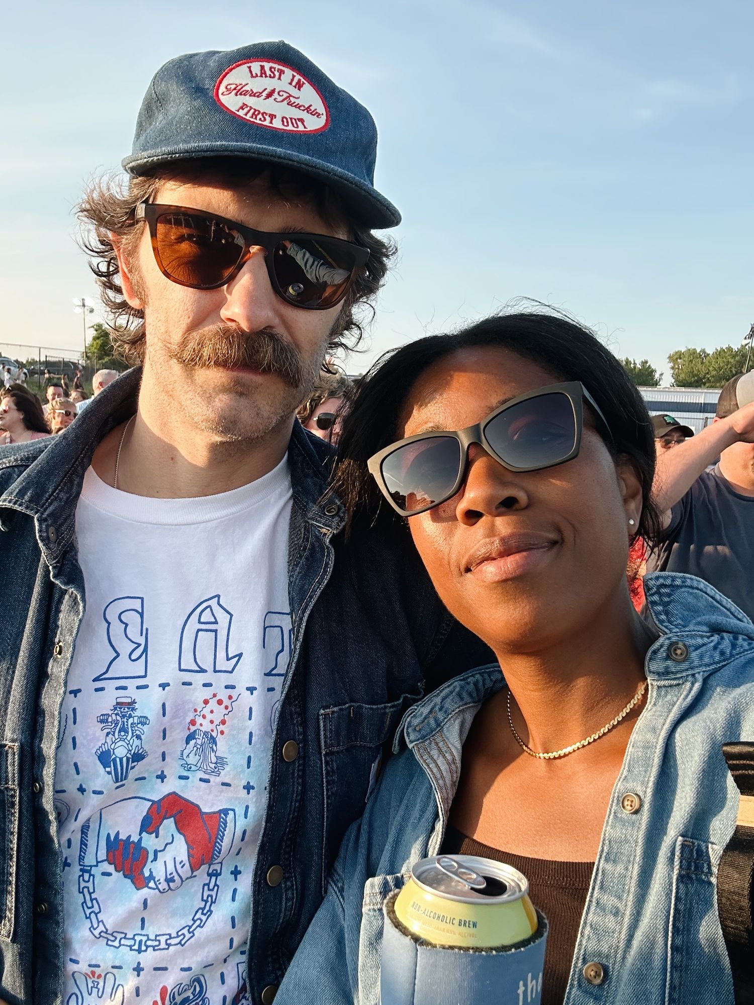 A man and woman wearing sunglasses and denim jackets pose together at an outdoor music festival.
