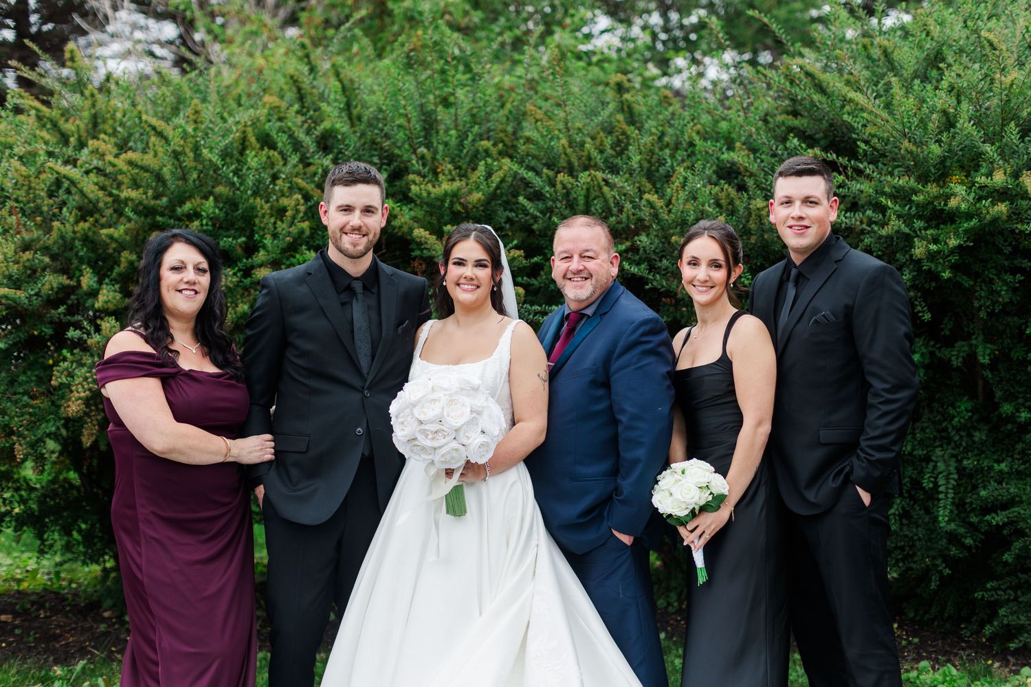 Bride in a white gown holding a bouquet poses outdoors with five family members in formal attire in front of green hedges.