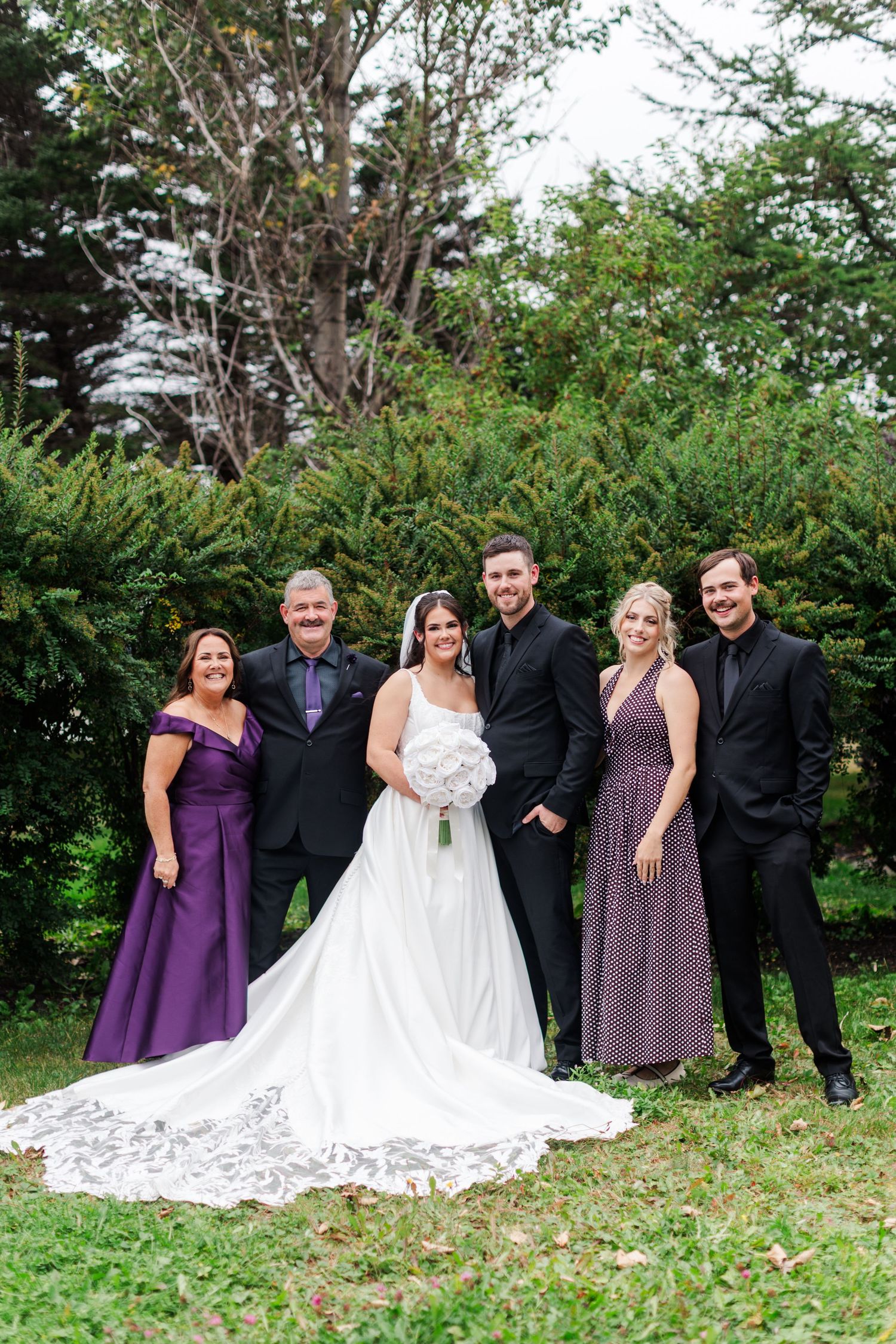 Bride in a white lace gown holding a bouquet poses outdoors on green grass with four family members in purple dresses and black suits.