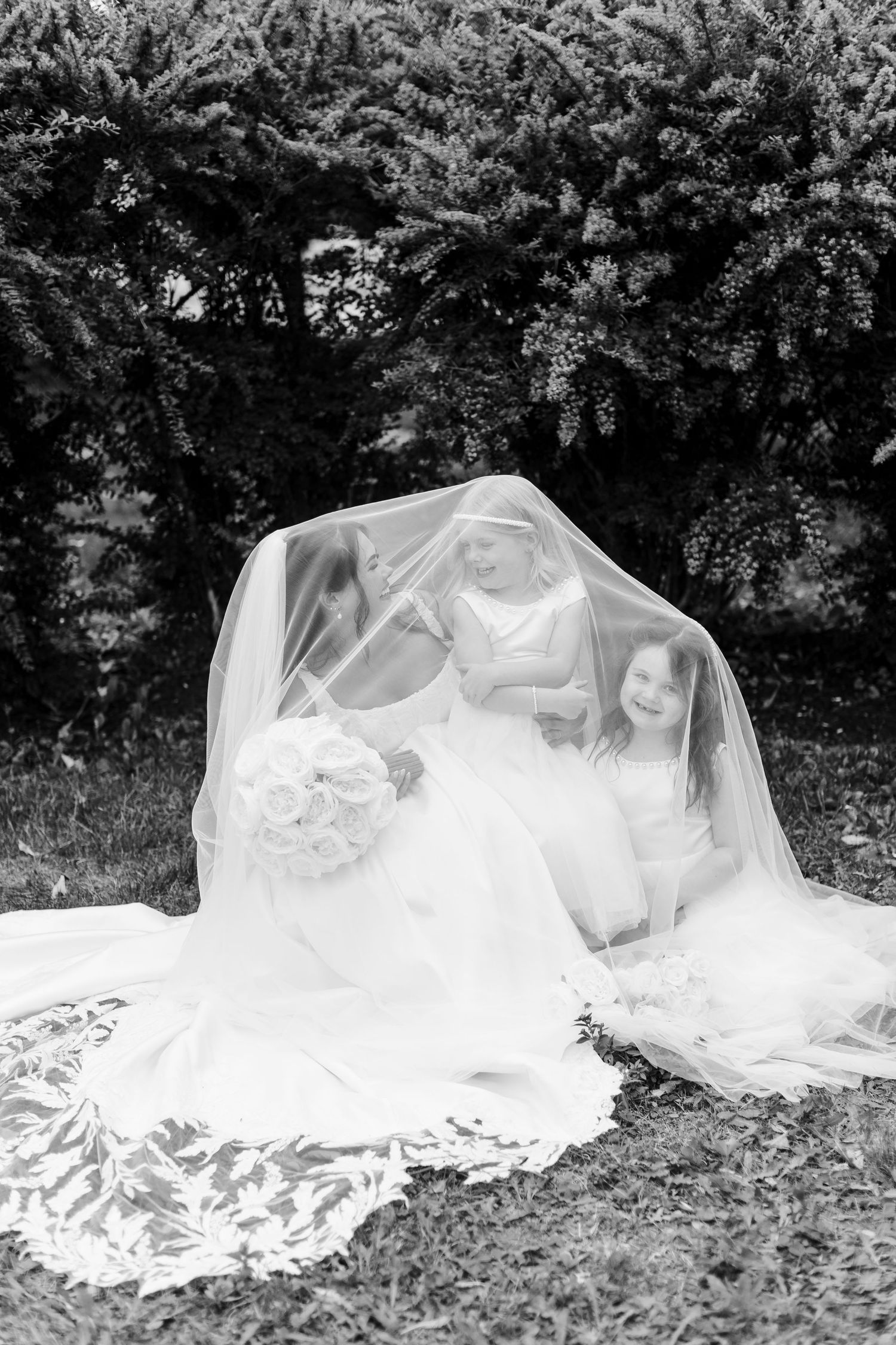 Black and white photo of a bride and two flower girls sitting on the grass together under her long lace-trimmed veil.