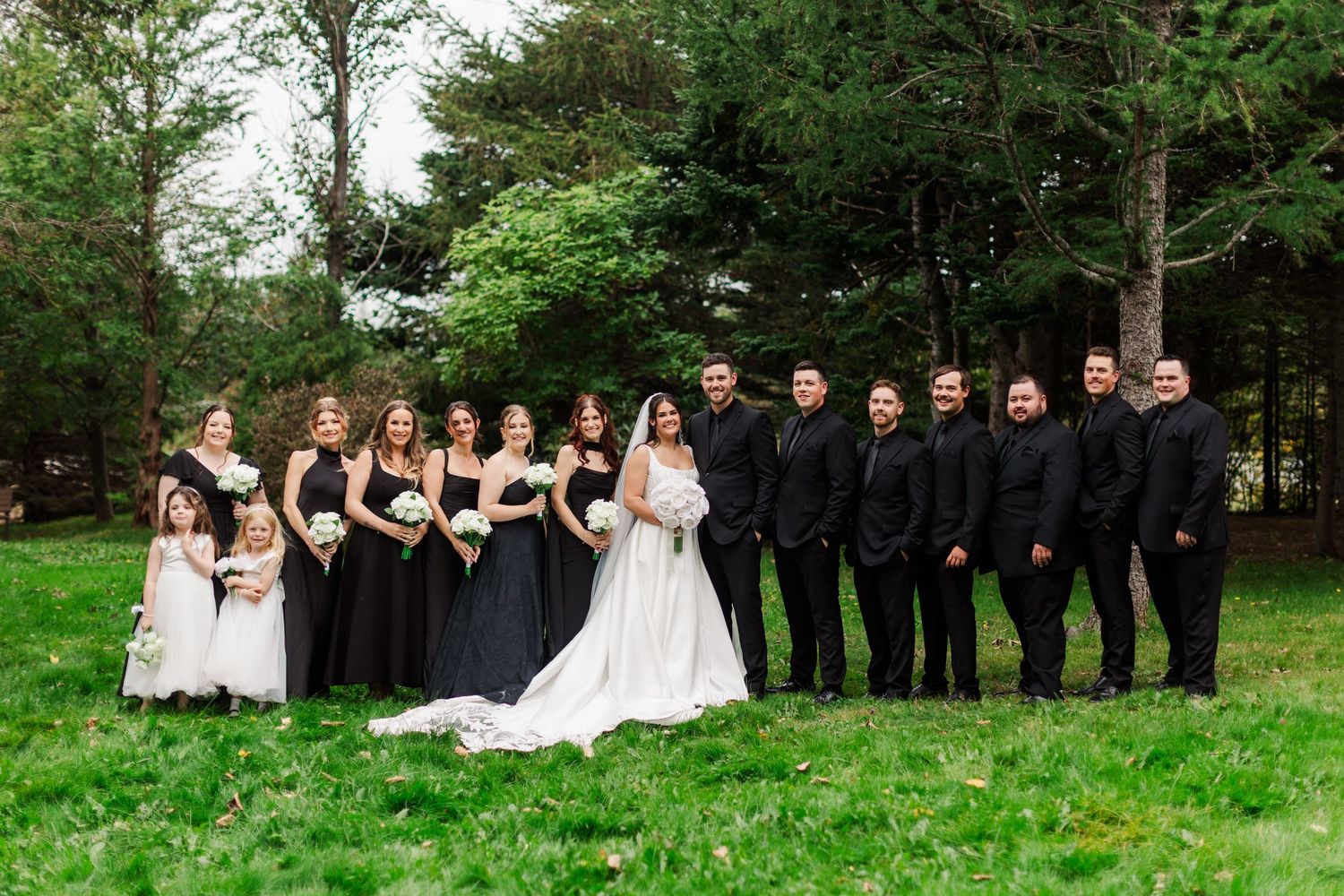 Bride in a white gown poses on a green lawn with bridesmaids in black dresses, flower girls, and groomsmen in black suits.