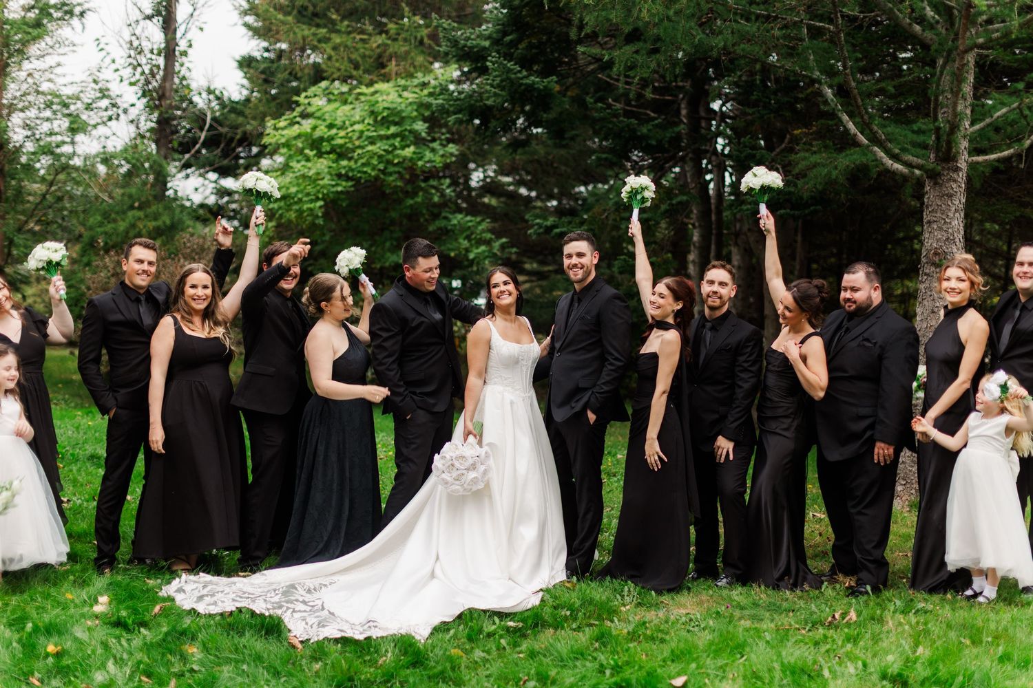 Bride in a white gown surrounded by groomsmen and bridesmaids in black, cheering with bouquets raised on a sunny green lawn.