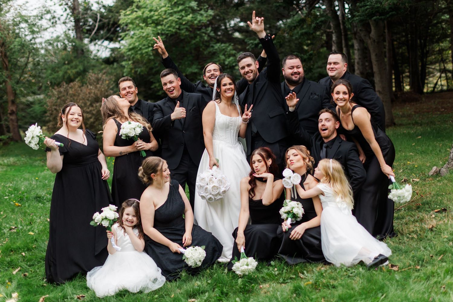 Joyful wedding party in black attire poses playfully on a grassy lawn with a bride in white and flower girls holding white bouquets.