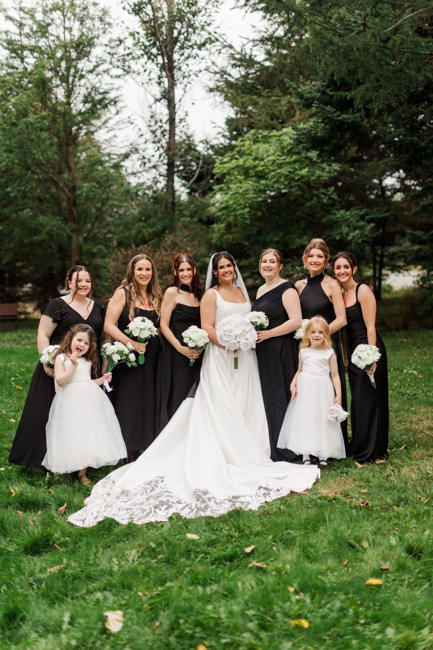 A bride in a white lace gown stands with six bridesmaids in black dresses and two flower girls on a grassy lawn.