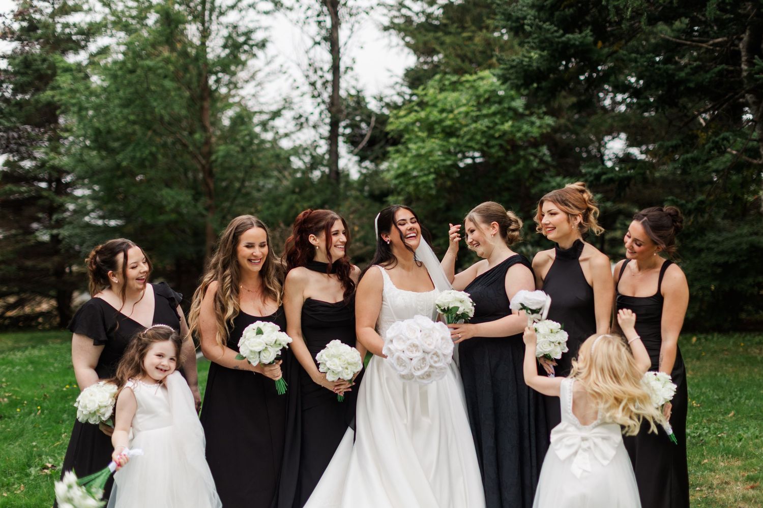 Bride in a white gown laughs with bridesmaids in black dresses holding white bouquets, with two flower girls beside them outdoors.