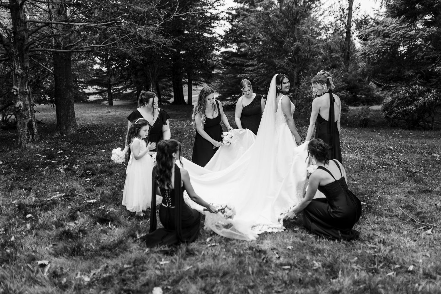 Black and white photo of bridesmaids in dark dresses kneeling to arrange the bride's long veil and gown beneath trees outdoors.
