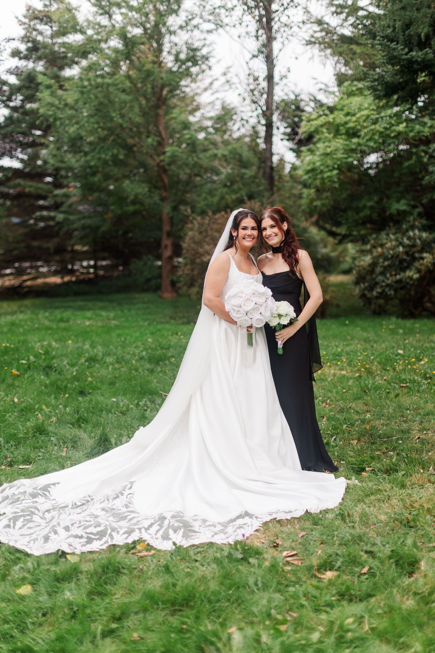 A bride in a white lace wedding gown with a long train poses with a bridesmaid in a black dress on a green lawn, holding white bouquets.
