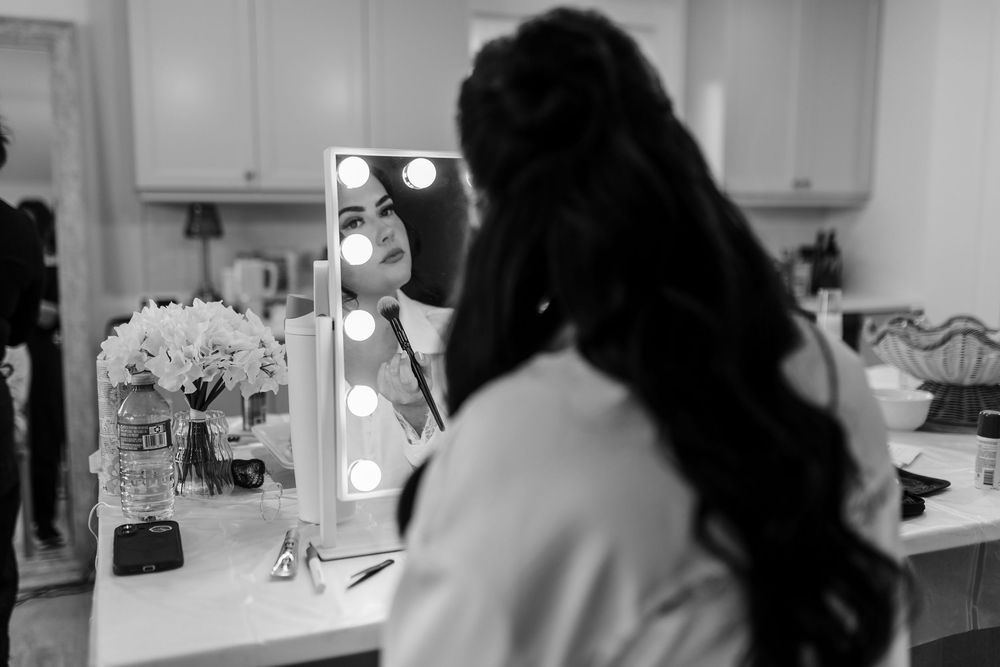 Black and white photo of a woman with long braids seen from behind, looking into a lighted vanity mirror at a makeup station with flowers.