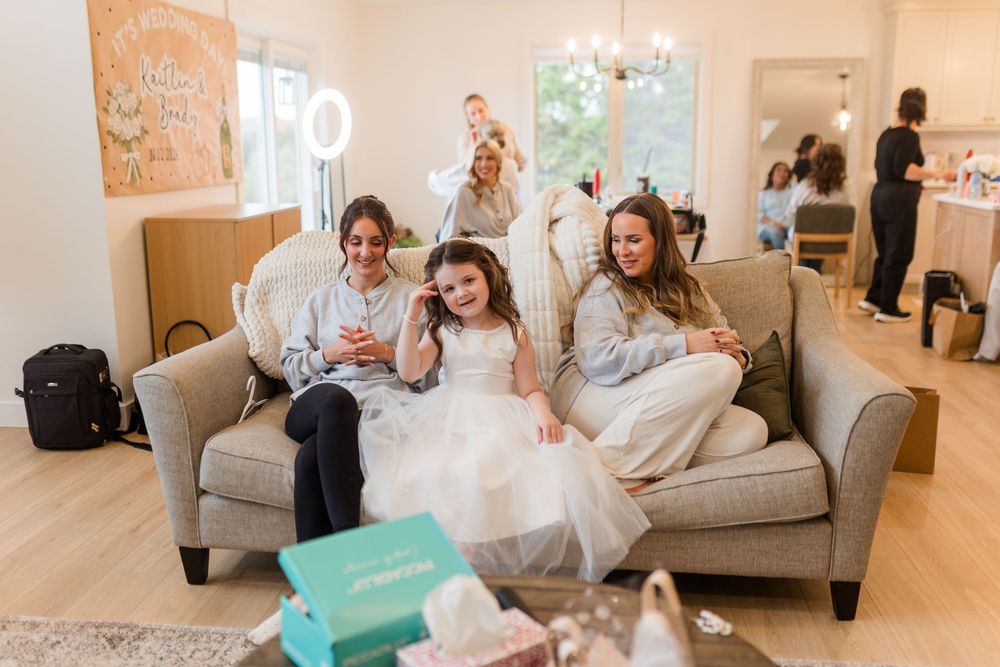 Young girl in a white tulle dress sits on a cream sofa between two smiling women in a bright living room with a teal gift box in front.