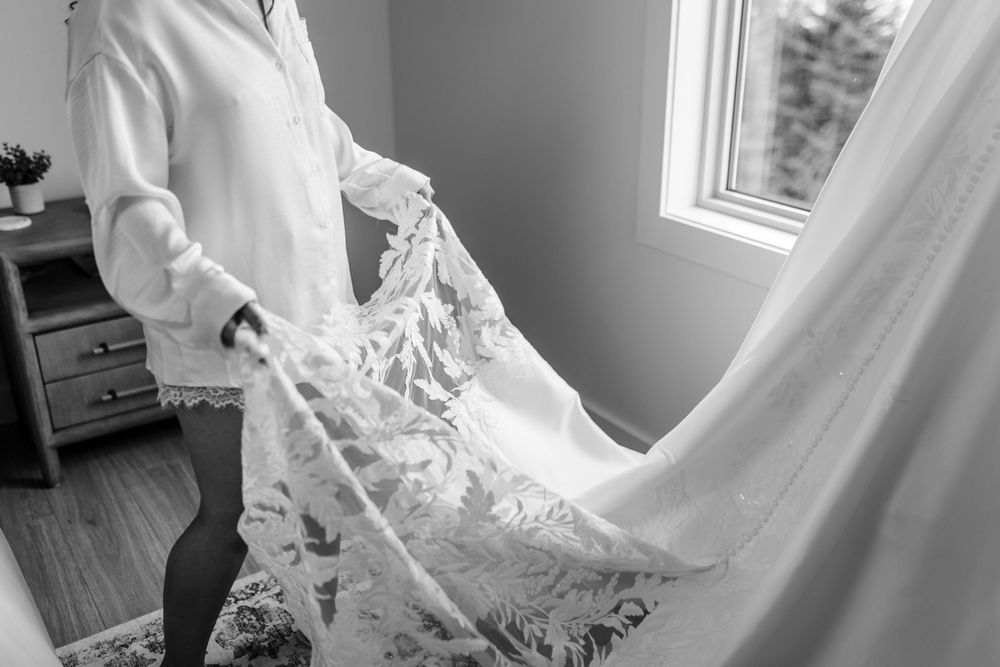 Woman in a white button-up shirt holds the delicate lace train of a wedding gown beside a sunlit window in a bright room.