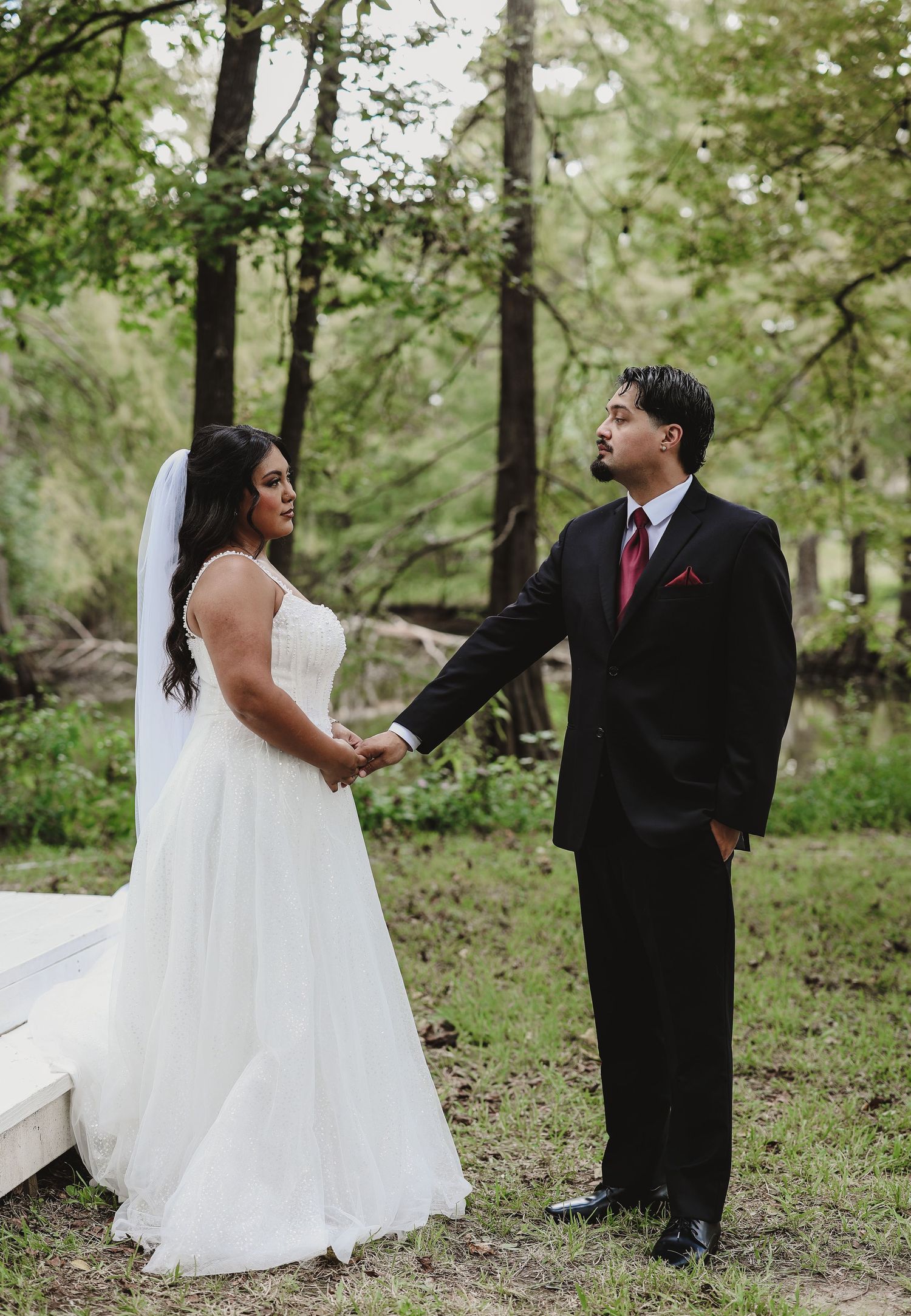 Bride in white gown and veil holds hands with groom in black suit and red tie outdoors in a wooded setting.