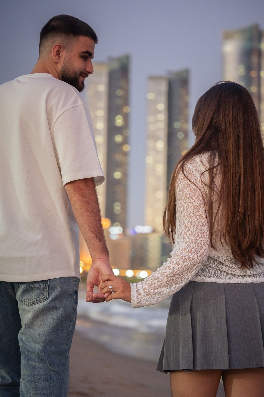 Couple holding hands on a beach at dusk with city skyline lights in the background.
