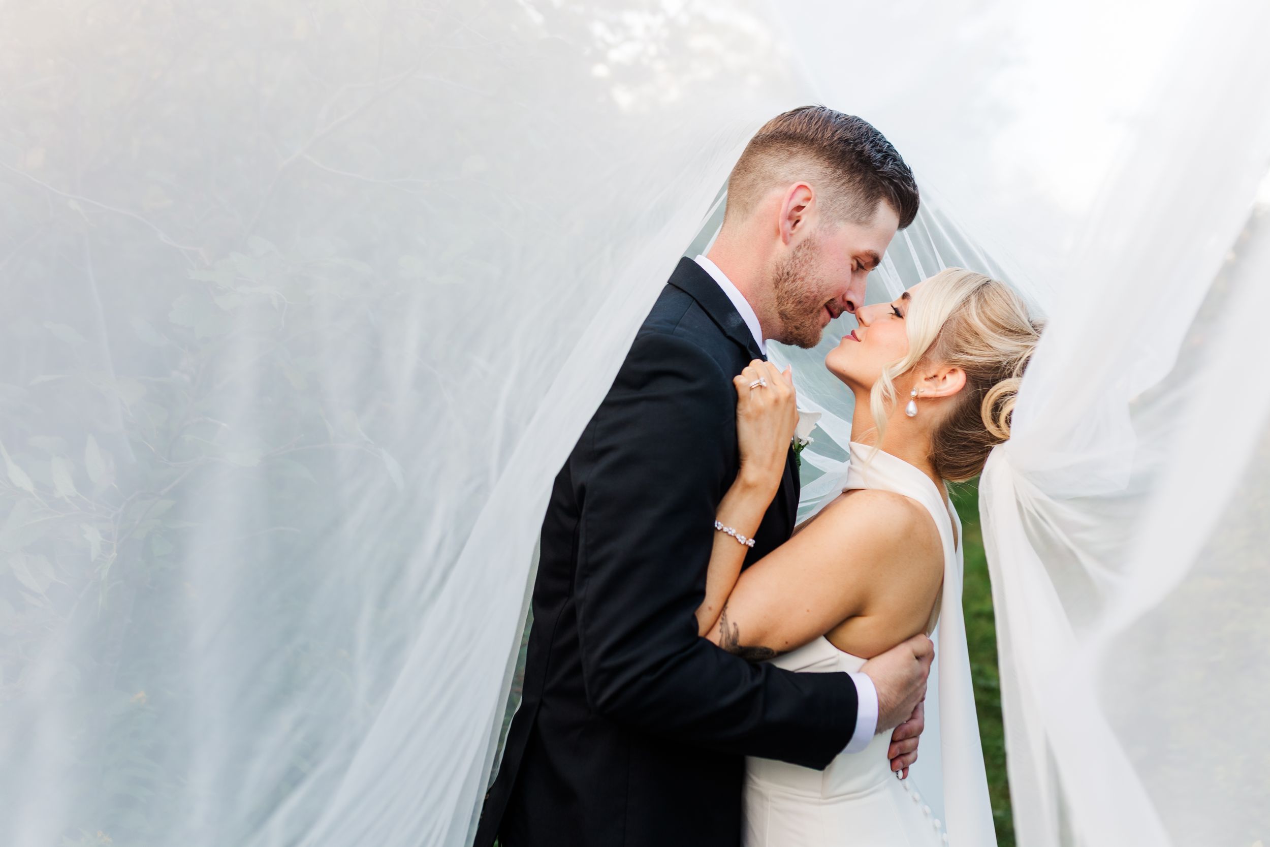 Bride and groom share a romantic moment under a flowing white veil outdoors on their wedding day.