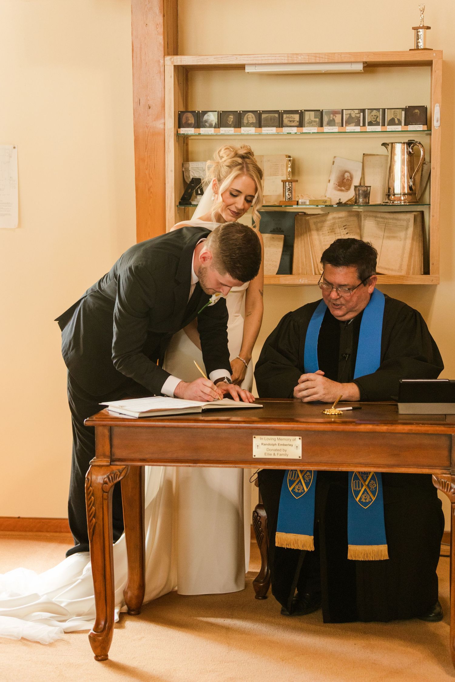 Bride and groom signing wedding registry with officiant in black robe at wooden desk during church ceremony.