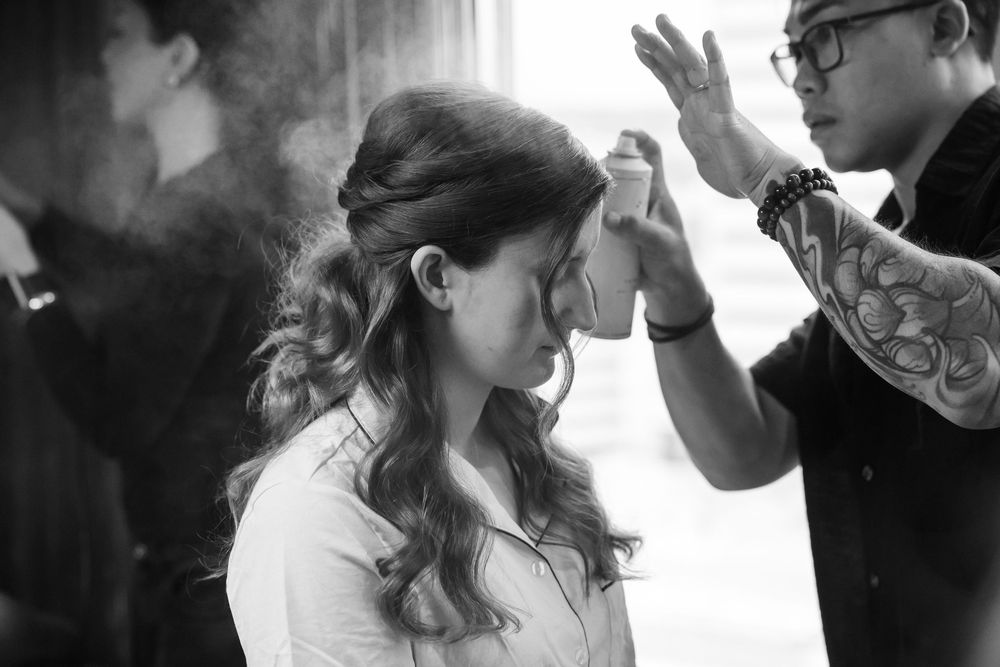 Black and white photo of a bride getting hair styled with spray by a tattooed hairstylist before her wedding.