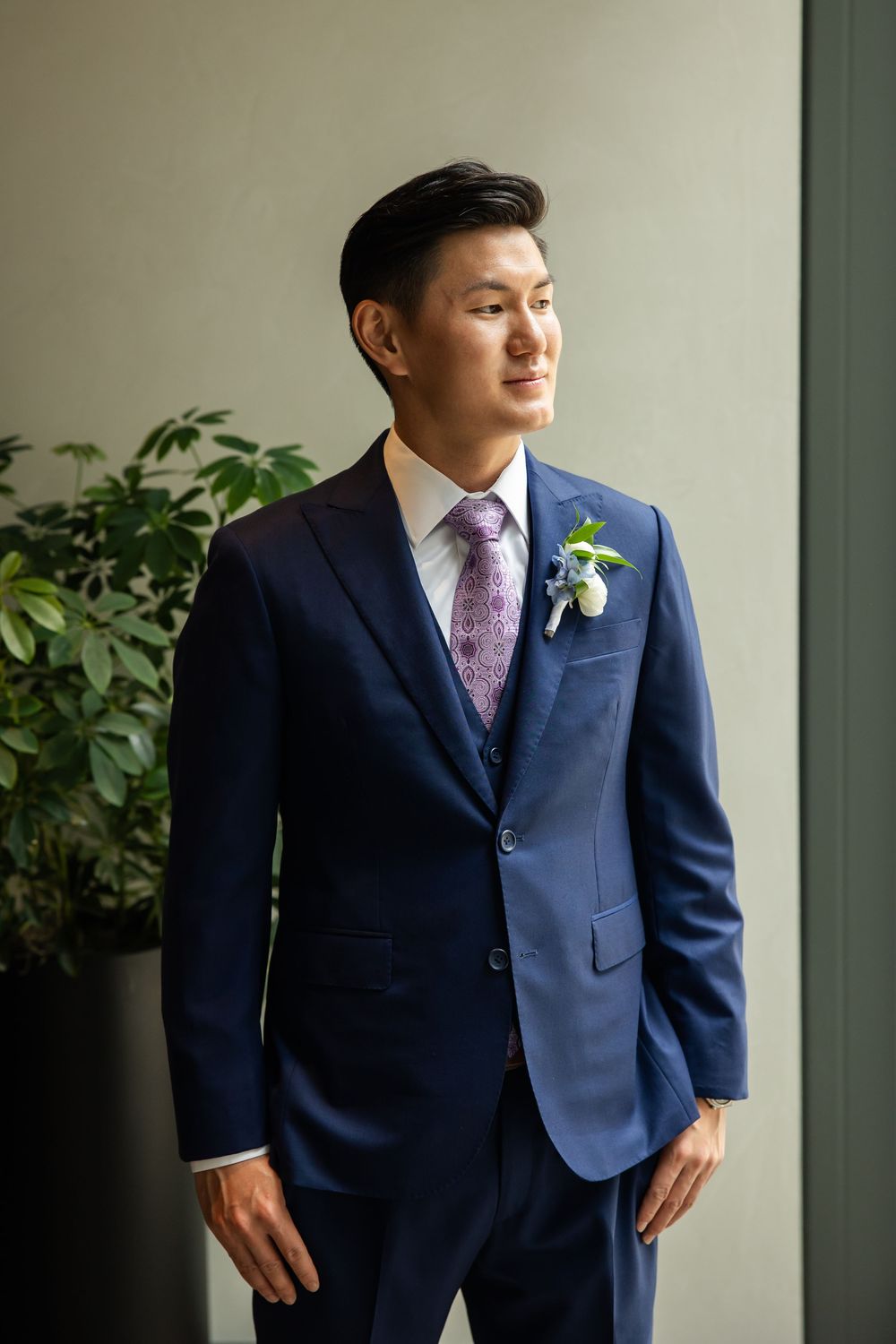 Groom in navy blue suit with purple tie and floral boutonniere poses elegantly indoors near green plants.
