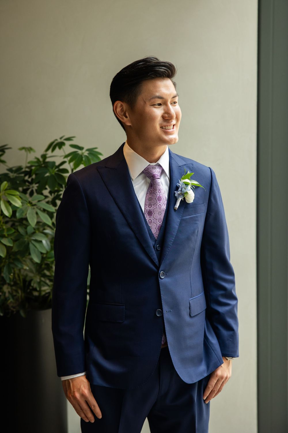 Smiling groom in navy blue suit with floral boutonniere and purple tie at wedding.