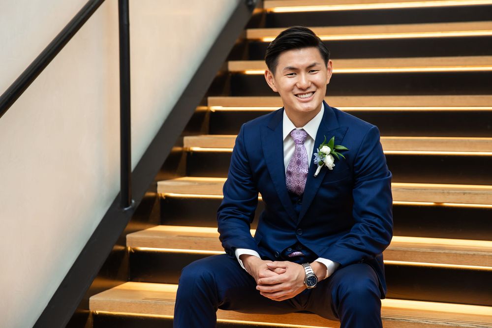 Smiling groom in navy suit with floral boutonniere sits on wooden stairs at wedding venue.