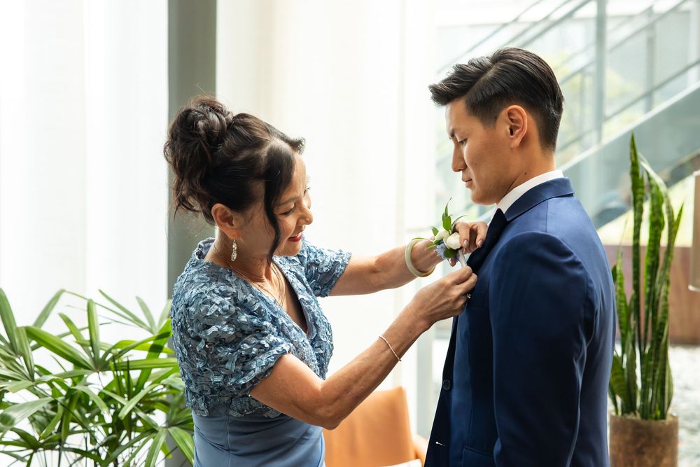 A woman pins a boutonniere on a man in a navy suit, surrounded by green plants in a bright indoor setting.