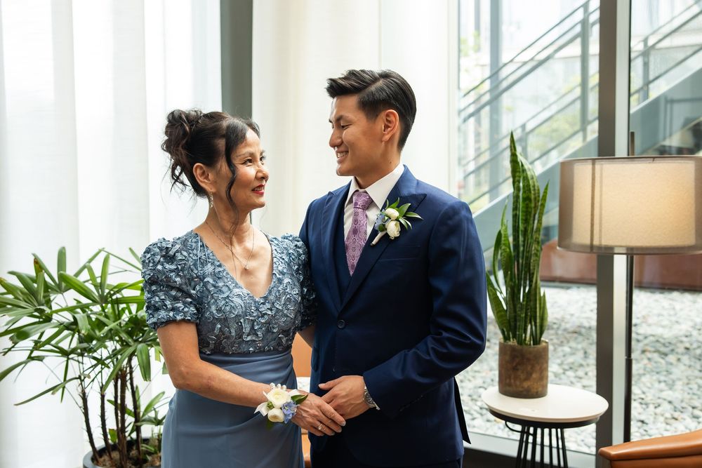 Couple in formal attire smiling at each other indoors, man in navy suit with boutonniere, woman in blue dress.